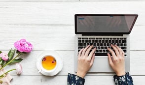 A woman with a blue flowery blouse is typing on her laptop keyboard. She has a cup of tea next to her and some pink flowers on her desk.